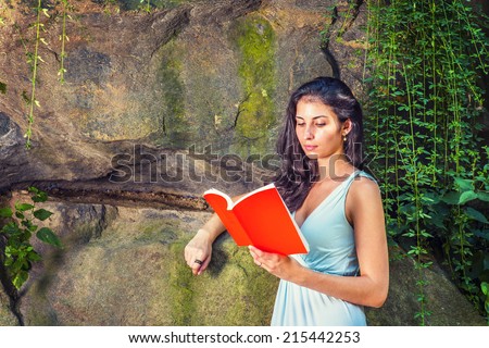 Pretty Lady Reading Outside. Dressing in a light blue dress, a young college student with long curly hair is standing by rocky wall with long green leaves, looking down, reading a red book.
