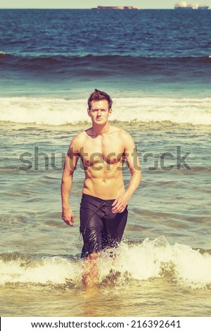 Portrait of Young Sexy Man. A well built young handsome muscular guy, half naked, wearing a bathing suit, is walking out of water on the beach, confidently looking forward. Sea Watching.