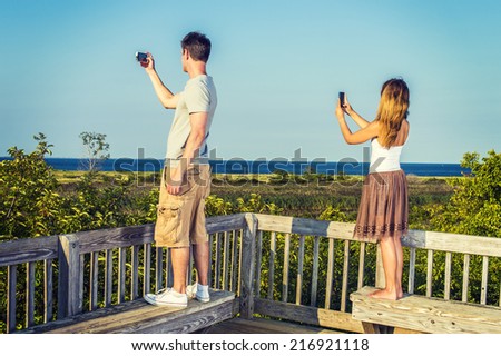Traveling couple video recording scenery of beach on remote location, using mobile phones, guy wearing gray T shirt, yellow pants, white sneakers, girl wearing golden brown skirt, white top, barefoot.