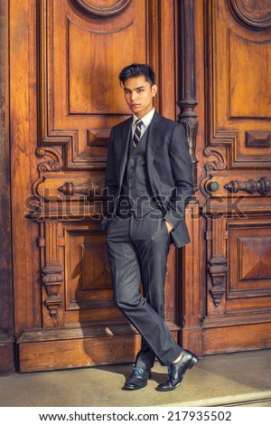 Business Man. Dressing in a black suit, patterned necktie, vest, leather shoes, hands in pockets, a young handsome college student is standing by an old fashion style office door, relaxing, thinking.