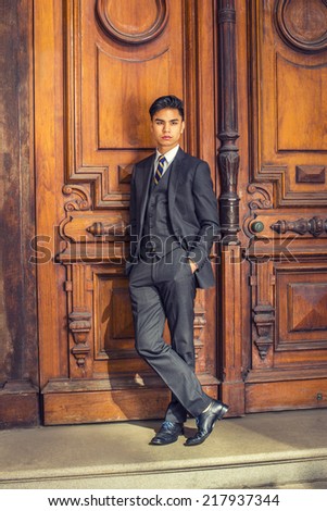 Business Man. Dressing in a black suit, patterned necktie, vest, leather shoes, hands in pockets, a young handsome college student is standing by an old fashion style office door, relaxing, thinking.