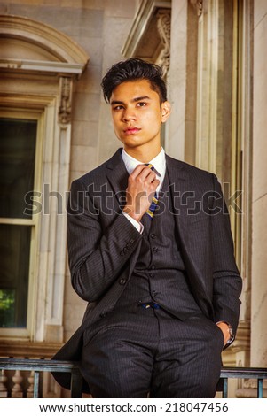 Business Man. Dressing in a black suit, patterned necktie, vest, a hand in pocket, a hand touching tie, a young handsome businessman is standing by a railing in office building, looking at you.
