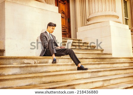 College Student Working Outside. Dressing in a black suit, patterned necktie, leather shoes, a young businessman is sitting on stairs outside an office, reading,  working on a laptop computer.