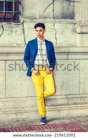 City Boy. Dressing in blue blazer, patterned under shirt, yellow pants, sneakers, hands on belt loops, rolled over cuffs, a young guy is standing against old style wall, bending a leg, looking at you.