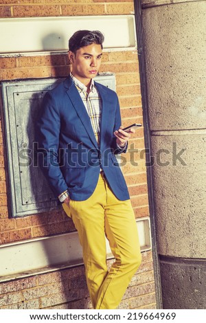 Waiting Your Phone Call. Dressing in blue blazer, yellow pants, hand holding a mobile phone.  a young handsome guy is standing by a brown brick wall, looking forward, lost in thought.