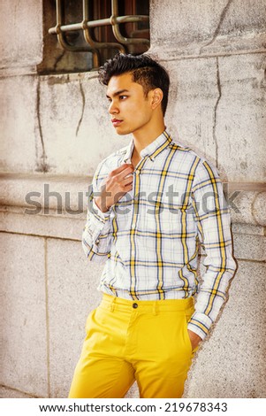 Man Thinking Outside. Wearing yellow and white, patterned shirt, yellow pants, a hands in pocket, a hand touching the collar,  a young handsome guy is standing by wall, lost in thought.
