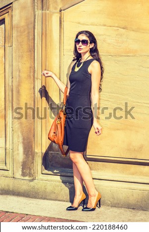 Business Lady. Dressing in black work dress, wearing sunglasses, necklace with golden pendant, leather shoes, arm carrying a brown bag, a young sexy businesswoman is standing outside, taking a break.
