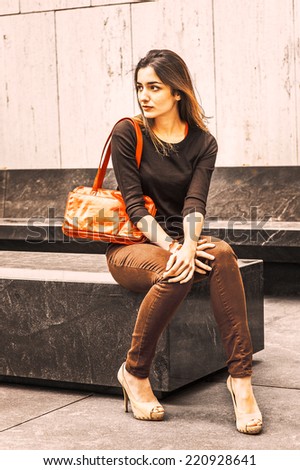 Professional Woman Relaxing. Wearing black long sleeve sweater, dark brown pants, shoulder carrying a red leather bag, a pretty lady is sitting on marble stone outside office building, relaxing.