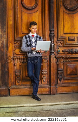 Man Working Outside. Wearing black, white, gray patterned sweater, red tie, blue jeans, leather shoes, a young guy is standing against an old style office door, looking down, working on a computer.