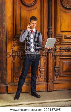 Man Working Outside. Wearing a patterned sweater, red tie, blue jeans, leather shoes, a young guy is standing outside office, talking on a mobile phone, working on a laptop computer in the same time.