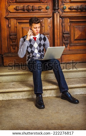 Man Working Hard. Wearing patterned sweater, red tie, blue jeans, leather shoes, a young guy is sitting on stairs by office door, talking on mobile phone, working on laptop computer in the same time.