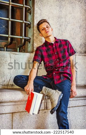 Man Relaxing Outside. Dressing in a short sleeve, black, red patterned shirt, jeans, boot shoes, a young handsome guy is sitting by a window, head leaning on wall, reading a red book, tired, relaxing.