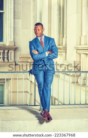 Dressing formally in blue suit, patterned undershirt, tie, leather shoes, short haircut, crossing arms and legs, a young black businessman is sitting on railing in office building, relaxing,