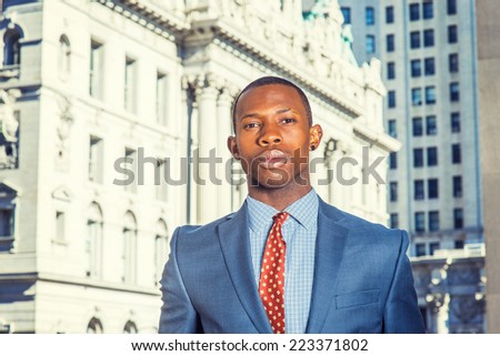 Portrait of Black Businessman. Dressing formally in blue suit, patterned undershirt, tie, short haircut, a young handsome black guy is standing in a business district, looking at you.