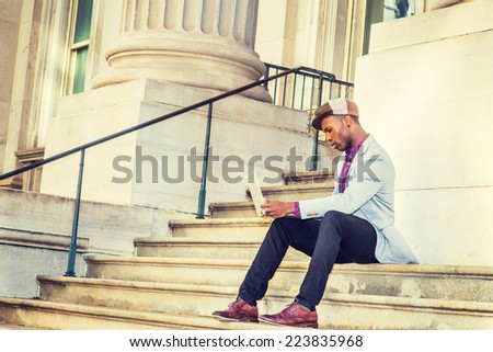 Man Working Outside. Wearing a Newsboy cap, dressing in light gray blazer, black pants, brown leather shoes, a young guy is sitting on stairs outside office building, working on laptop computer.