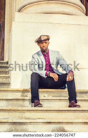 Man Urban Fashion. Wearing a Newsboy cap, dressing in light gray blazer, patterned pink, black under shirt, black pants, brown leather shoes, a young guy is sitting on stairs, relaxing.