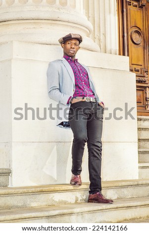 Man Urban Fashion. Wearing a  Newsboy cap, dressing in light gray blazer, patterned pink, black under shirt, black pants, brown leather shoes, a young guy is leaning back on a column, relaxing.