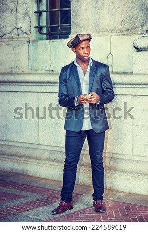 Man Urban Fashion on Street. Wearing fashionable Newsboy cap, dressing in black blazer, pants, brown leather shoes, a young black guy is standing by the wall with a window, waiting for you. Texting.