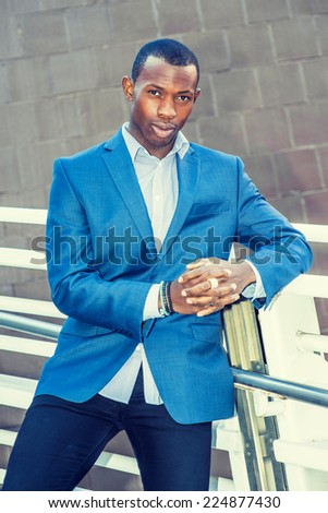 Portrait of Young Business Man. Wearing a blue blazer, black pants,, short haircut, bracelets, finger ring, a young black guy is standing against metal fences by the wall, confidently looking at you.