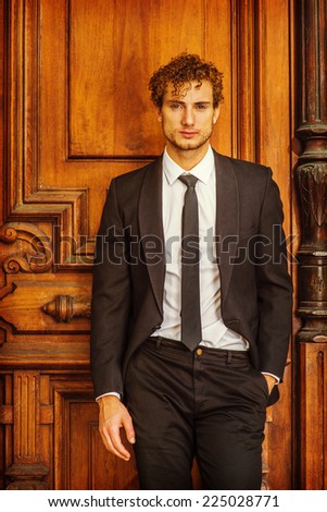 Portrait of Sexy Classic Business Man. Dressing in black suit with Shawl Lapel, unbuttoned, black necktie, white under shirt, a young handsome guy with golden curly hair casually standing by door way.
