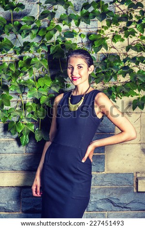 Pretty Lady. Dressing in black work dress, a necklace with golden pendant, hand resting on hips, a young sexy businesswoman is standing outside against the wall with green ivy leaves, looking at you.