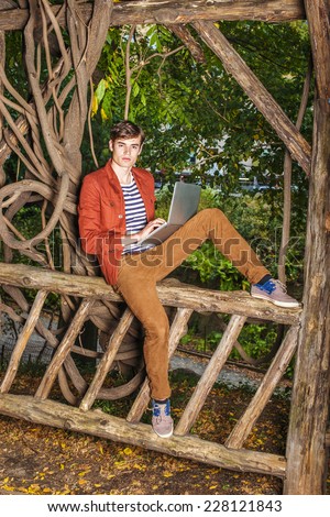 Man working outside. Wearing a dark reddish brown jacket, unbuttoned, brown corduroy pants, a young college student sitting on a wooden fence with rattan trees, working on laptop computer, thinking.