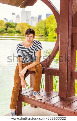 Man Waiting for You. Wearing a striped T shirt, brown corduroy pants, a young handsome guy is standing by a lake, bending over, looking at you, relaxing. Big city high building scene in background.