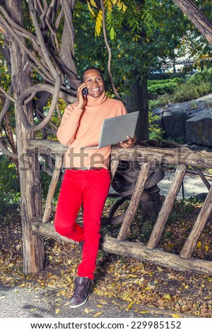 Man Working Outside. Dressing in light orange sweater with high collar, red pants, patterned boot shoes, a young black guy is standing by fence, smiling, making phone call, working on laptop computer.