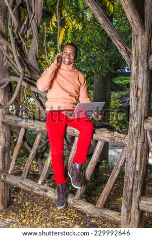 Man Working Outside. Dressing in light orange sweater with high collar, red pants, patterned boot shoes, a young black guy is sitting on fence, smiling, making phone call, working on laptop computer.