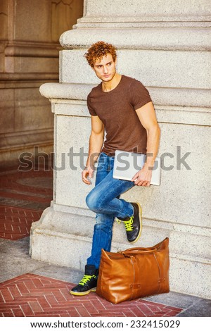 Traveler Relaxing on Street. Wearing dark brown T shirt, jeans, sneakers, a leather bag on ground. a young sexy guy with curly hair is standing against wall, holding laptop computer, waiting for you.
