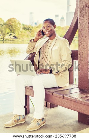 Business Man Working. Dressing in cream blazer, collarless sweater, white pants, sneakers, a young black guy is sitting by a lake in big city, talking on phone, smiling, working on laptop computer.