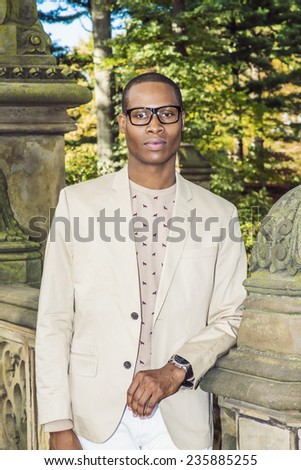 Portrait of Young Black Professor. Dressing in cream blazer, collarless sweater, wearing eye glasses, wristwatch, A young black graduate student is standing on campus, seriously looking at you.