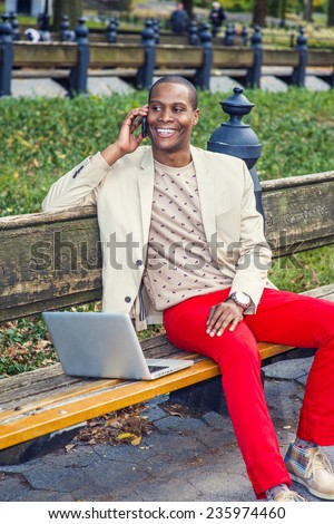 Man Working Outside. Dressing in cream blazer, collarless sweater, red pants, sneakers, a young black guy is sitting on the bench, smiling, making phone call, working on laptop computer. Modern Life.