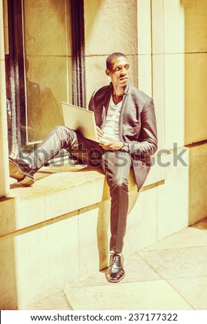 Man Working Outside. Dressing in fashionable jacket, pants, leather shoes, a young black college student is sitting against a window frame, looking up, reading, thinking, working on a laptop computer.