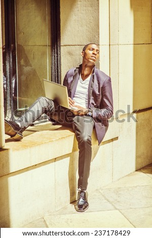 Man Working Outside. Dressing in fashionable jacket, pants, leather shoes, a young black college student is sitting against a window frame, looking up, sad, thinking, working on a laptop computer.