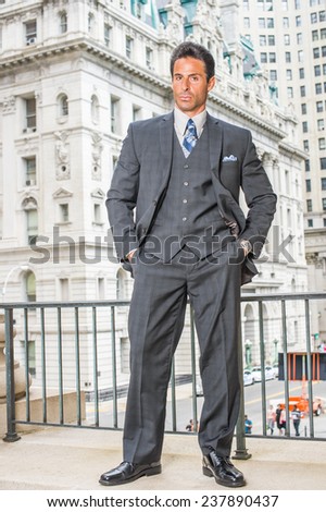 Man Power. Dressing in dark blue three piece suit, necktie, leather shoes, hands in pockets, a handsome, sexy, middle age businessman is standing outside office building, confidently looking at you.