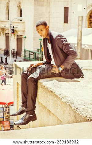 Young man thinking on street. A young black college student is sitting outside, arched the back, under strong sunshine, looking down, sad, lost in thought.