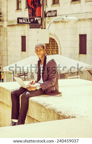 Young man working on street. A young black college student is sitting outside, looking away, thinking, working on a laptop computer. Wall Street sign in the background.