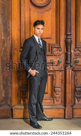 Dressing formally in three pieces black suit, patterned necktie, vest, leather shoes, hands in pockets, a young handsome businessman is standing by an old fashion style office door, looking at you.