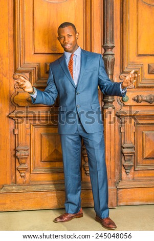 Dressing formally in blue suit, patterned shirt, necktie, leather shoes, short haircut, wearing ear stud, a young guy is standing in the front of old style office door, opening arms, welcoming you.