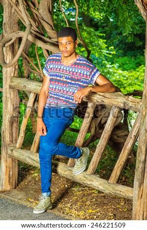 Young Black Man Waiting for You. Wearing a colorful pattern shirt, blue jeans, sneakers, necklace, a young handsome guy is standing against a wooden fence, charmingly looking at you.