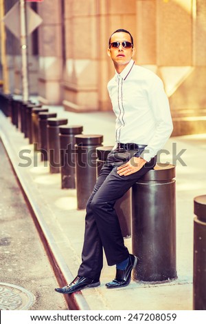 Man Street Fashion. Dressing in a white shirt, black pants, leather shoes, wearing sunglasses, a young, mysterious guy is sitting on the street, relaxing and waiting for you. Instagram filtered look.