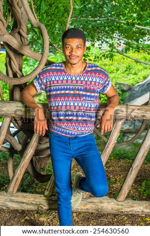Portrait of Young Black Man. Wearing a colorful pattern shirt, blue jeans, necklace, a young handsome guy is standing against a wooden fence, smiling, charmingly looking at you.