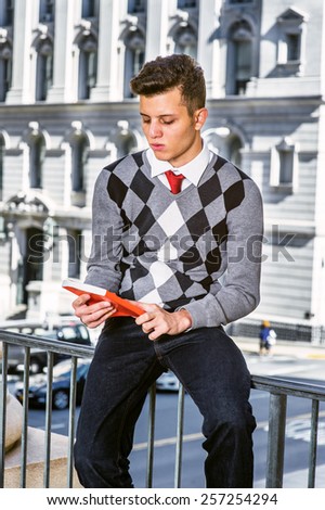 Man Reading Outside.  Dressing in black, white, gray patterned sweater, jeans, a young college student sitting on a railing outside, holding red book, looking at the cover. Instagram filtered look.