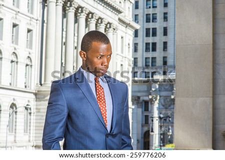 Businessman thinking hard. Dressing in blue suit, patterned tie, a young black guy standing in the front of vintage style office building, looking down, frowned, confused, sad, lost in thought.