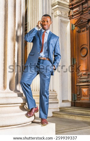 Dressing in blue suit, patterned tie, leather shoes, a young black businessman standing outside vintage style office building, smiling, calling on his mobile phone during a working break.