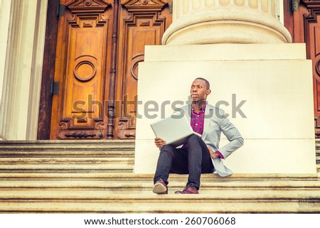 Working outside. Dressing in light gray blazer, black pants, brown leather shoes, a young black guy is sitting on stairs outside vintage style office building, hands holding laptop computer, thinking.