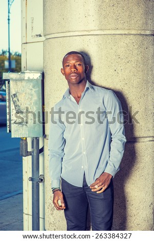 Man Waiting for You. Wearing a long sleeve shirt, black pants, short haircut, a young black guy standing against column with metal box and pipe line, relaxing, taking a break. Instagram effect..