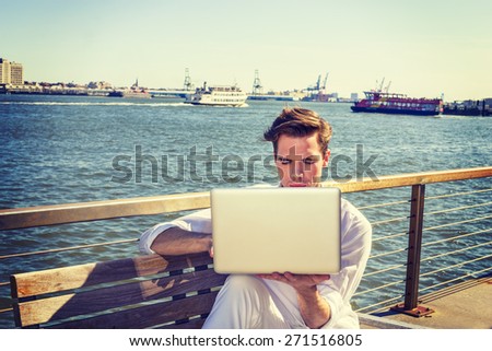 Study Outside. Young blonde, handsome student, wearing white shirt, sitting by river, reading, working on laptop computer. small boats, big city on far background. Traveler.