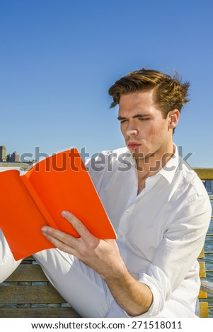Power of Reading. Young blonde, handsome student, dressing in white, hands holding a red book, looking down, reading, thinking, under blue sky,